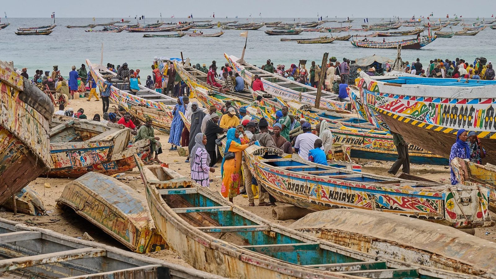 Painted fishing pirogues drawn up on a crowded beach, with more boats anchored offshore